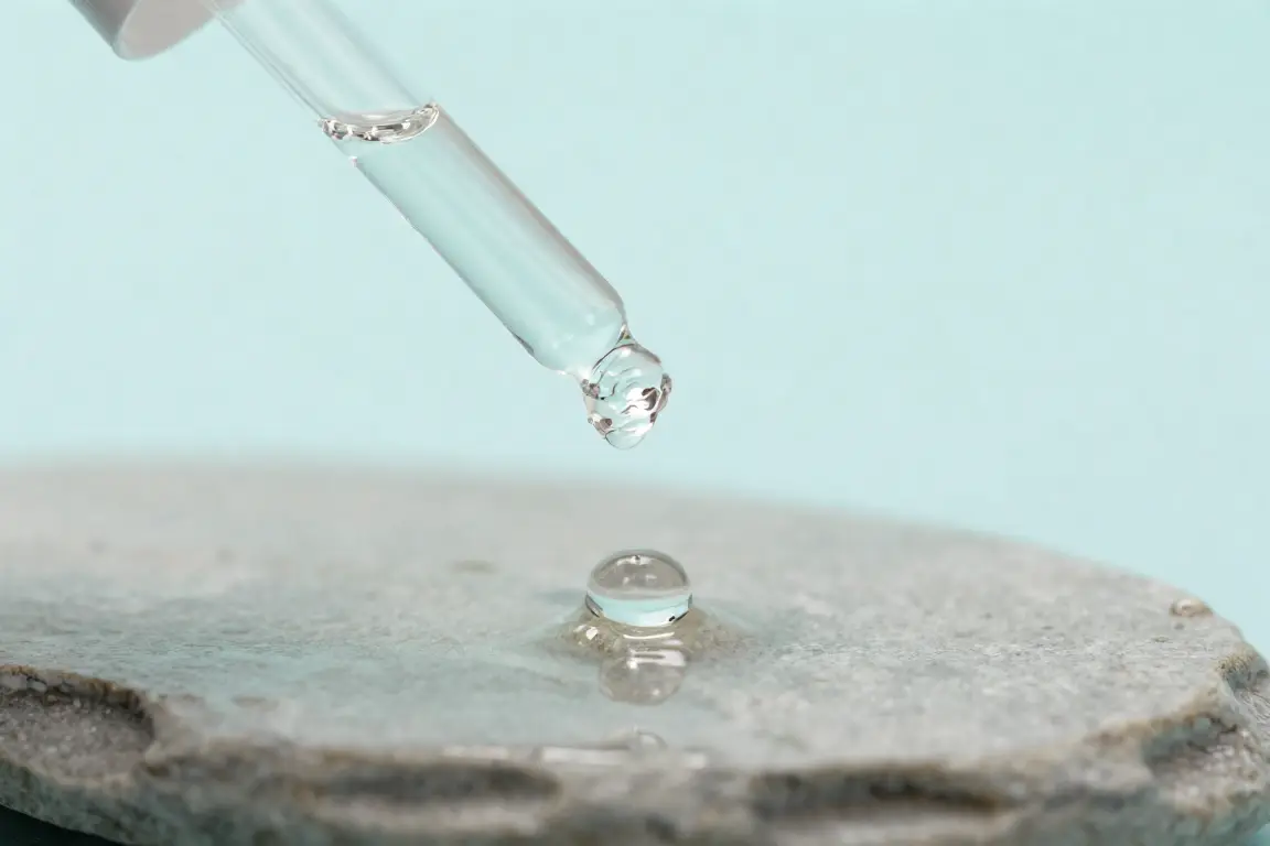 Glass dropper bottle dispensing a dewy hyaluronic acid serum droplet onto a wet stone surface.